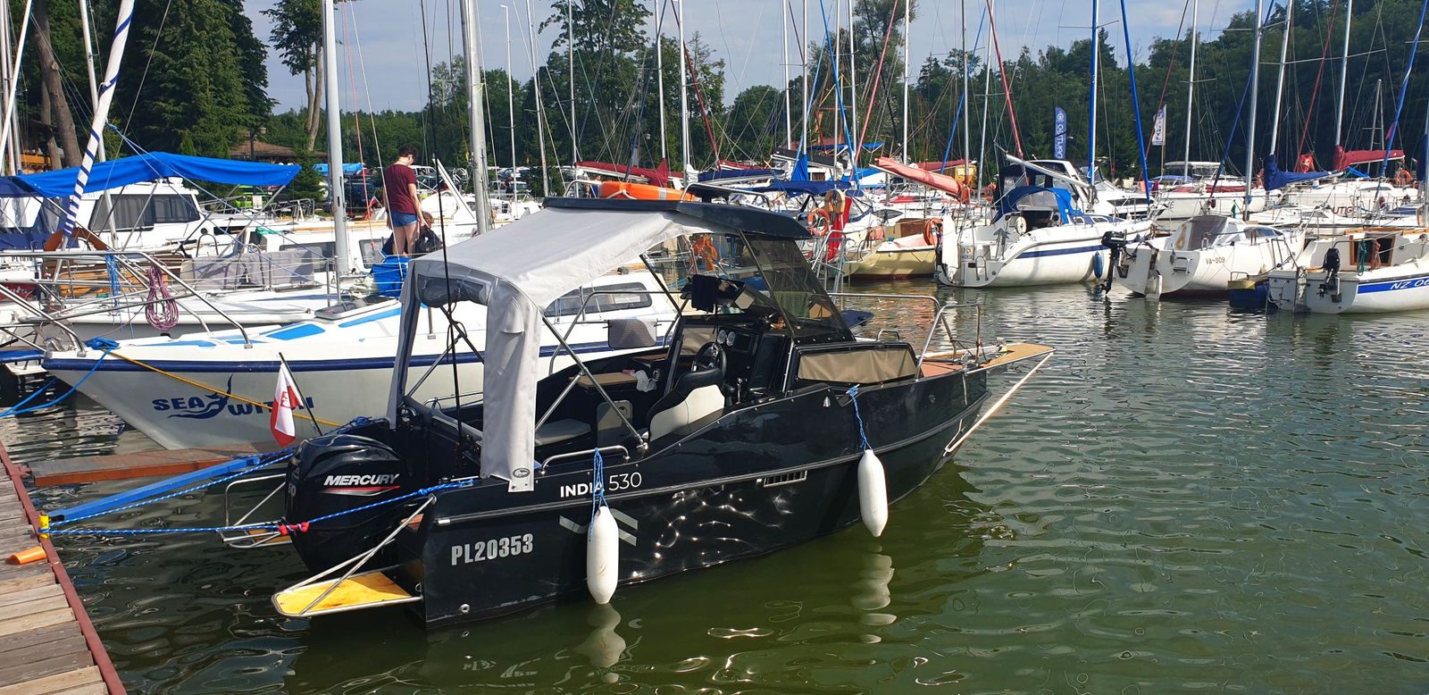 Ciemna łódź motorowa z daszkiem bimini przy drewnianym pomoście w marinie, na rufie silnik Mercury
