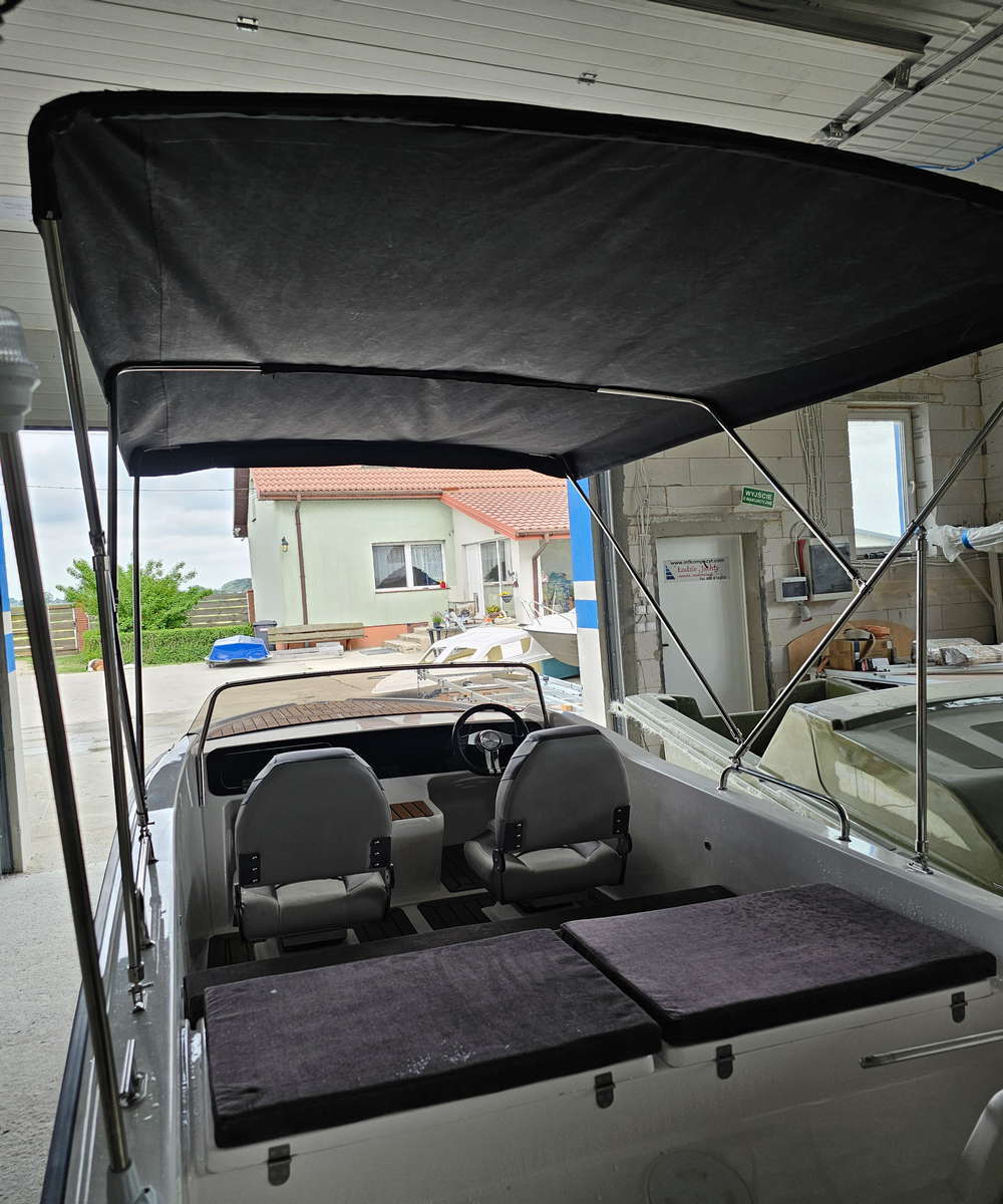 Kokpit łodzi motorowej INDIA CLASSIC z rozłożonym czarnym dachem bimini, dwoma fotelami i kierownicą, widok od rufy w stronę dziobu w hali stoczni